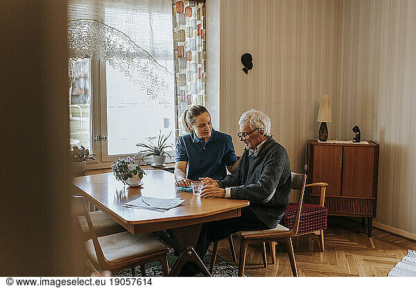 Weibliche Pflegehelferin im Gespräch mit einem älteren Mann bei der Einnahme von Medikamenten zu Hause
