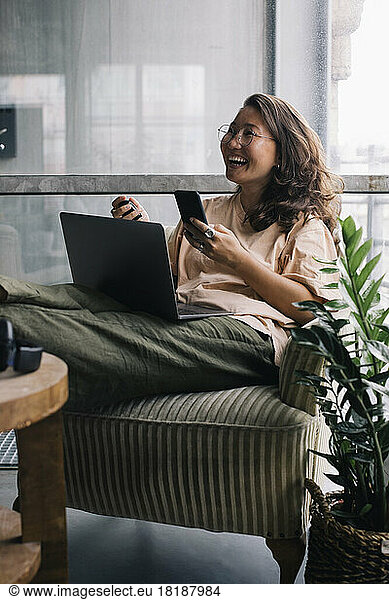 Weiblicher Computerprogrammierer mit Laptop und Smartphone lachend im Büro