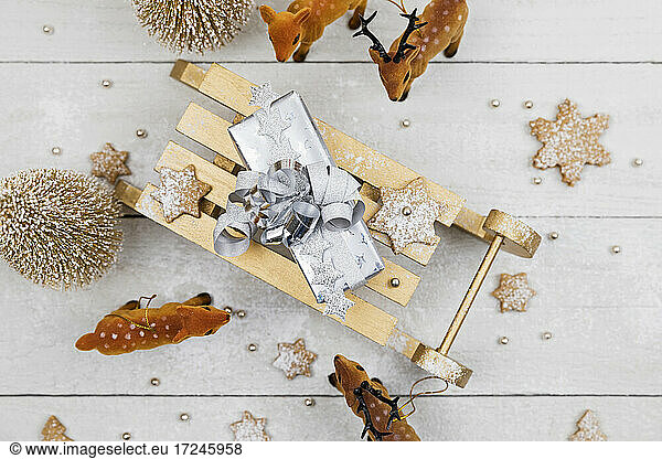 Weihnachtsgeschenk auf Spielzeug-Schlitten  umgeben von Spielzeug-Hirschen mit selbstgebackenen Keksen vor weißem Holzhintergrund