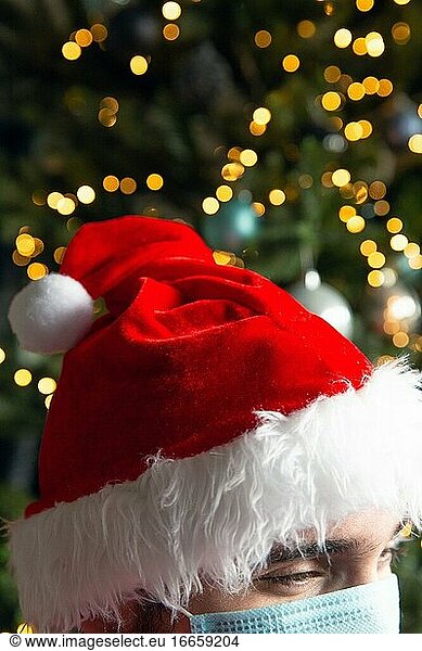 Weihnachtsmann Mütze mit Weihnachtsbaum auf dem Hintergrund Bokeh Lichter  trägt medizinische Maske für Covid-19  Weihnachten und Coronavirus Konzept Nahaufnahme.