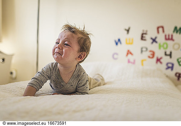 Weinendes Baby auf Bett mit weißer Decke und Alphabet an der Wand hinter ihm