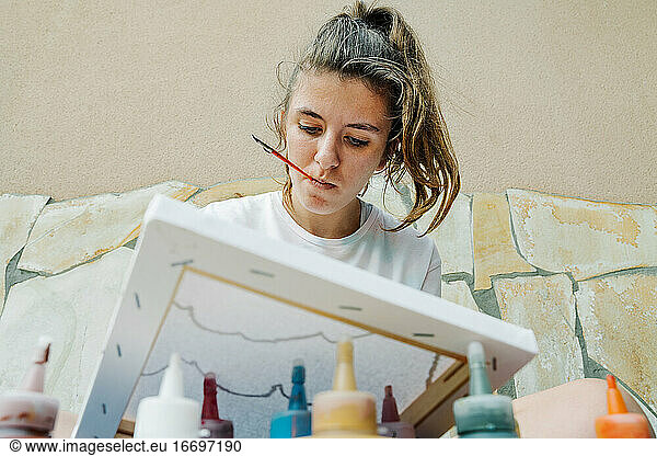 white girl biting a bush while painting with her finger her picture in front of bottle paintings. Overhead horizontal photo