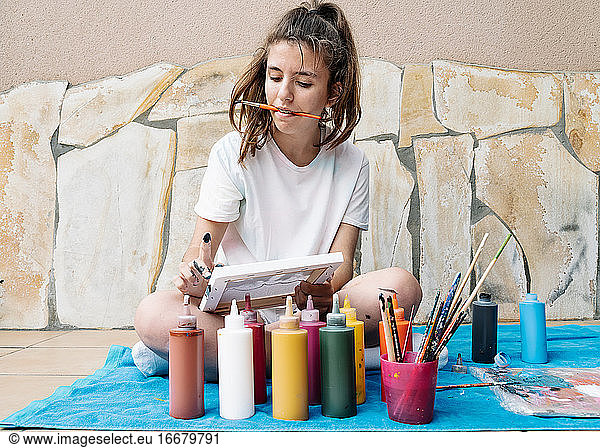 white girl biting a painting brush while painting with her finger her picture siting on a terrace in front of bottle paintings. Horizontal photo