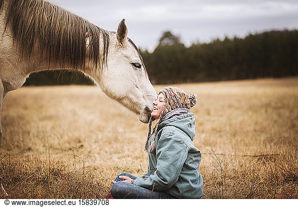 white horse tugging on girls hat in field in the fall while girl sits