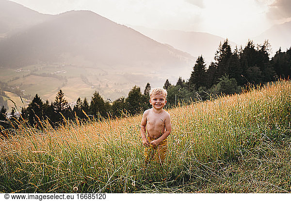 White male toddler smiling standing topless in the grass in mountains