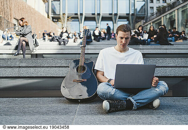 white man using laptop computer in square beside his classical guitar
