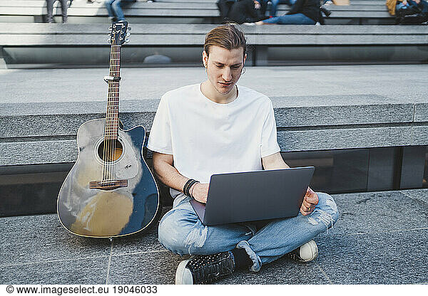 white man using laptop computer in square beside his classical guitar