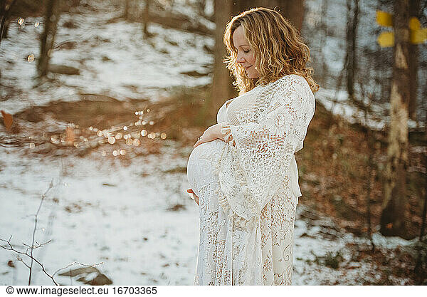 White pregnant mom smiling looking at her belly on sunny day in woods