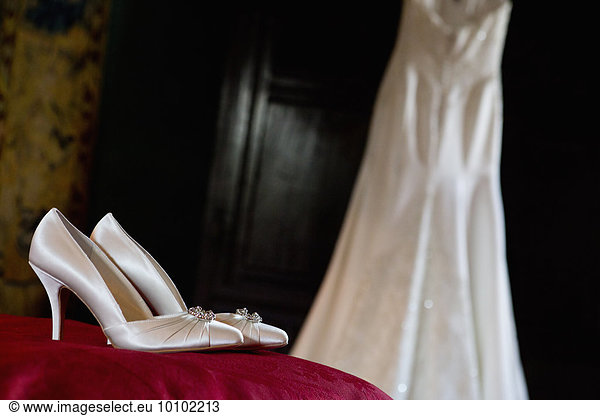 White satin high heeled wedding shoes and a dress hanging up.