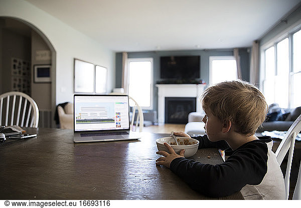 Wide Side View of Blonde Boy Eating Cereal Watching Lesson on Laptop