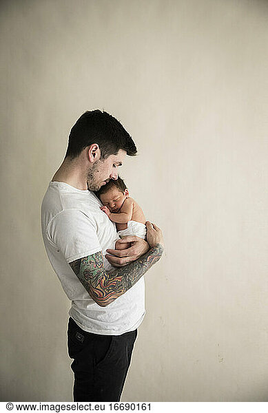 Wide Side View of Tattooed Millennial Dad Snuggling Diapered Newborn