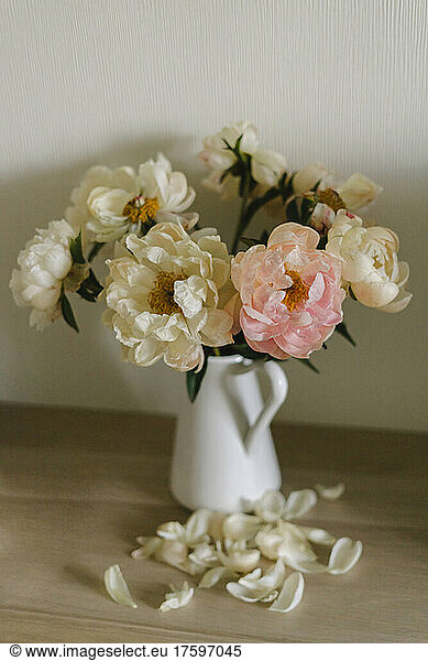 Wilted peonies in vase by petals on table