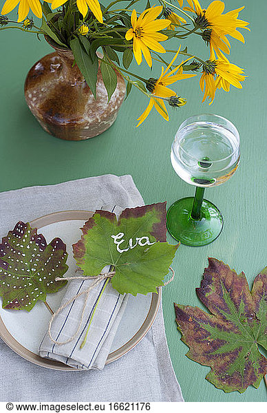 Wineglass with water  vase with blooming Jerusalem artichoke flowers and name tag made of autumn leaf