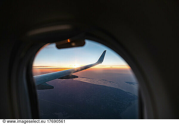 Wing of airplane flying against setting sun seen through porthole