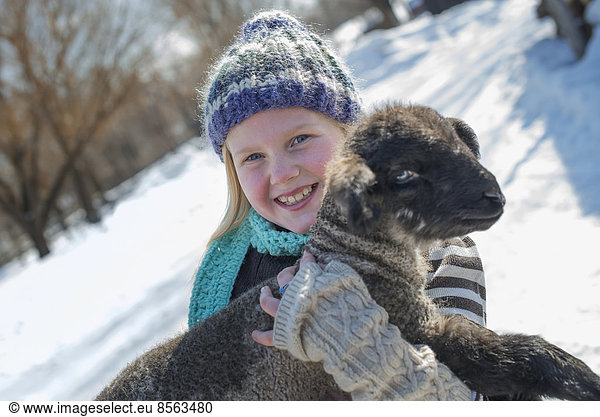 Winterlandschaft mit Schnee auf dem Boden. Ein junges Mädchen hält ein junges Lamm.
