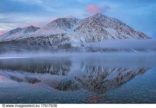 Winterliche Landschaft mit schneebedeckten Bergen  die sich im stillen Wasser des Kathleen Lake spiegeln  mit nebligen  tief hängenden Wolken und Pastellfarben  die eine stimmungsvolle Atmosphäre schaffen; Haines Junction  Yukon  Kanada