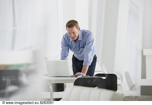 Wirtschaft. Ein Mann  der über einem Schreibtisch steht und sich herunterlehnt  um einen Laptop-Computer zu benutzen.