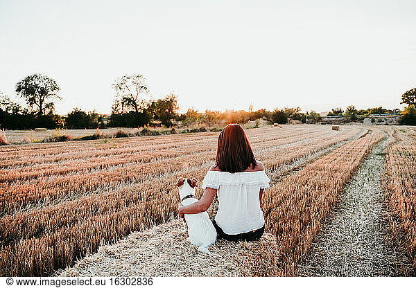 Woman and dog sitting on straw bale during sunset