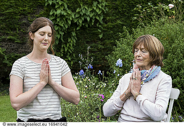 Woman and female therapist seated in a garden  hands together and eyes closed.