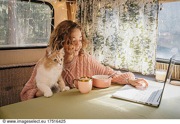 Woman and ginger cat in trailer are eating breakfast at laptop.