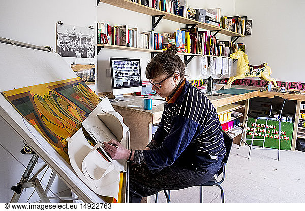 Woman artist sitting at a tilted drawing table in her studio  drawing  using several sheets of stiff paper.