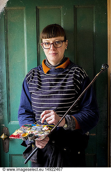 Woman artist wearing glasses holding paintbrush  palette and Maulstick  looking at camera.