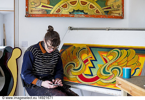 Woman artist working sitting by a drawing in progress at drawing table in a workshop  checking her mobile phone.