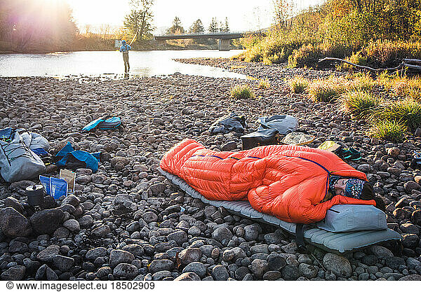 Woman asleep in sleeping bag on river edge with fiisherman behind