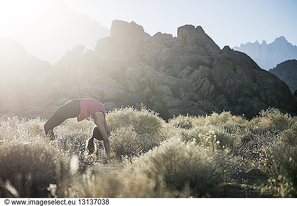 Woman bending over backwards while practicing yoga on field against mountains during sunny day