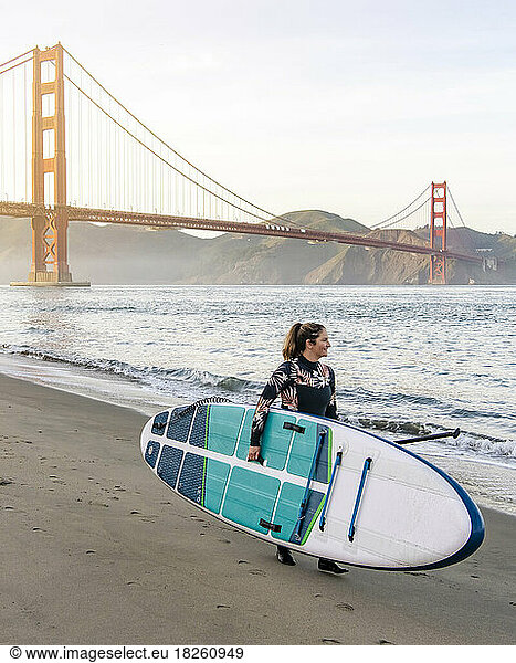Woman carrying paddleboard walking towards Golden Gate Bridge