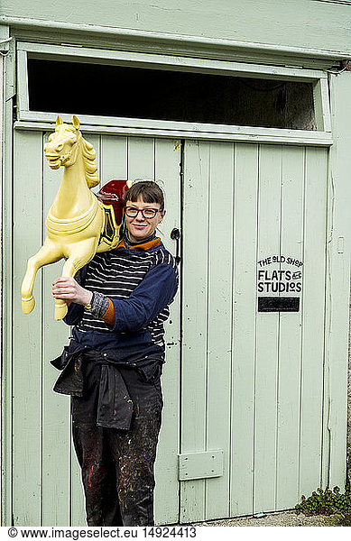 Woman carrying traditional wooden painted carousel galloper horse from merry-go-round on her shoulder outside a workshop