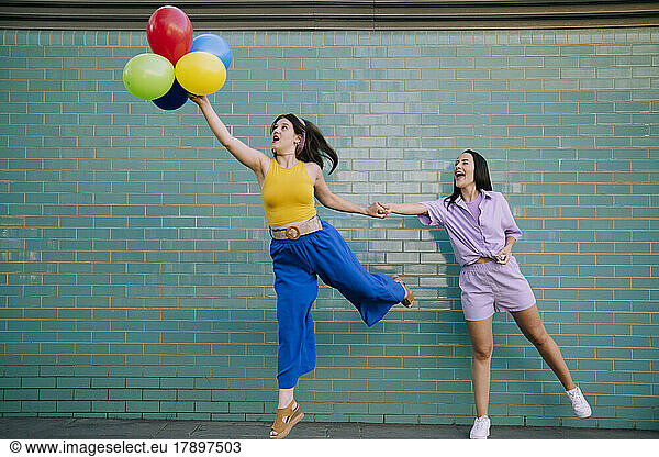 Woman catching friend holding multi colored balloons in front of turquoise wall