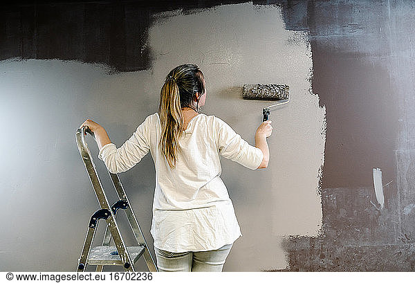 Woman climbed a ladder and catching a paint roller full of grey painting on a brown wall. Painter is upping and downing roller covering the wall with grey painting what remains wet. Horizonta