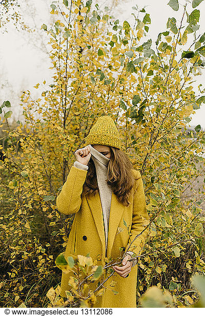 Woman covering face while standing against plants during autumn