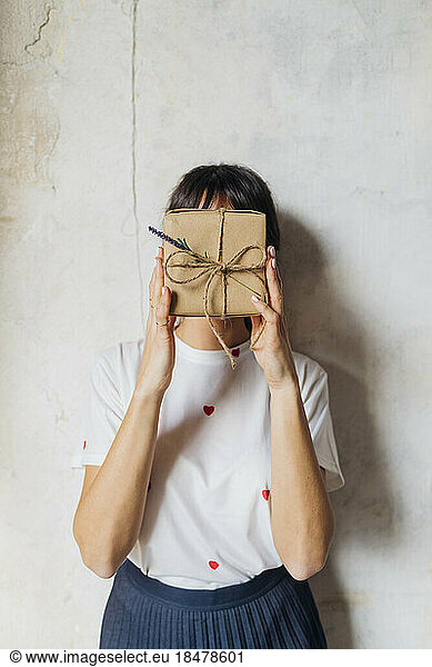 Woman covering face with gift box in front of wall