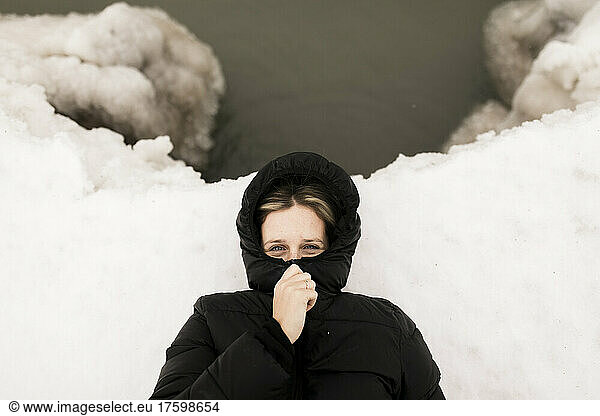 Woman covering face with hooded sweater lying down on snow in winter