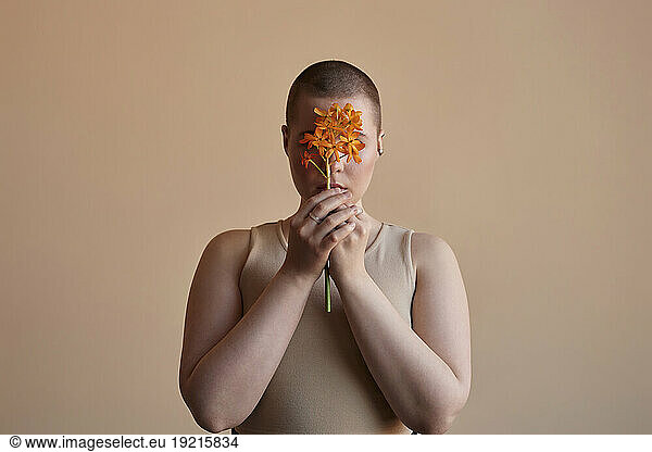 Woman covering face with orange flower against beige background