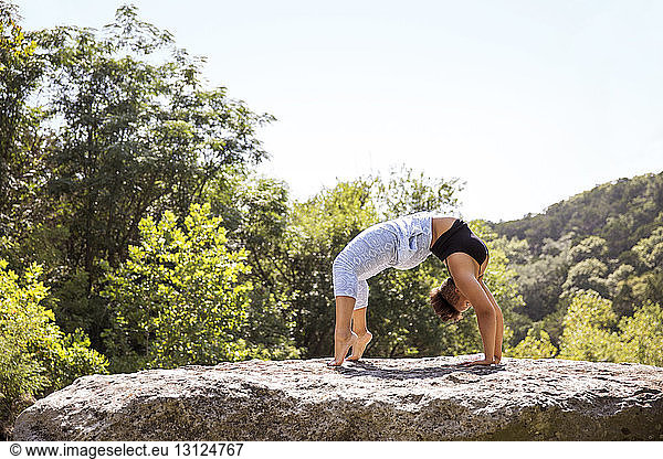 Woman doing bridge pose on rock against clear sky at forest