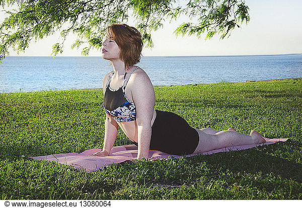 Woman doing cobra pose on exercise mat at park against sea and clear sky