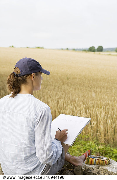 Woman drawing on paper while sitting in front of farm against clear sky