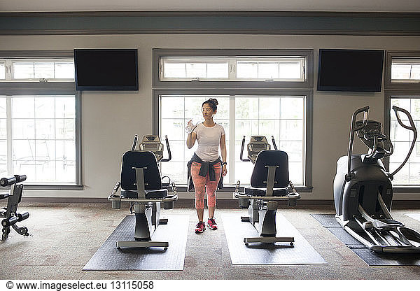 Woman drinking water while standing amidst machines at gym