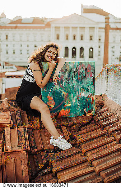 Woman embracing her artwork with happy face while sitting on tile roof