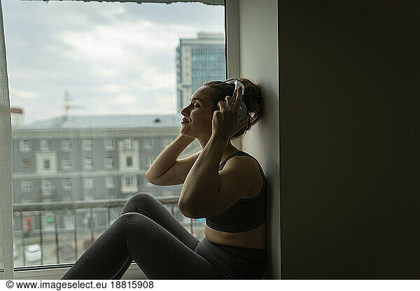 Woman enjoying music with headphones sitting on window sill at home