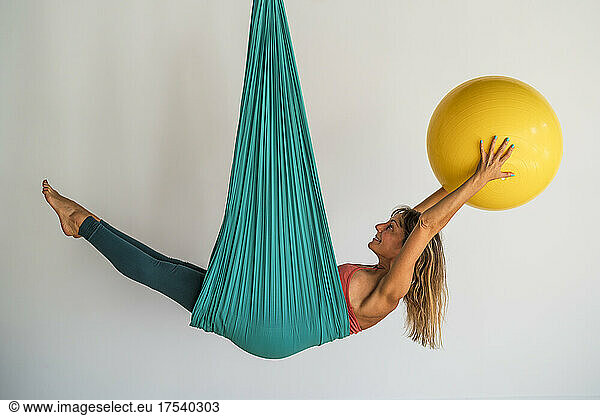 Woman exercising with fitness ball hanging on aerial silk in front of wall