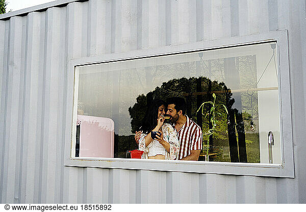 Woman feeding boyfriend in kitchen seen through window of container home