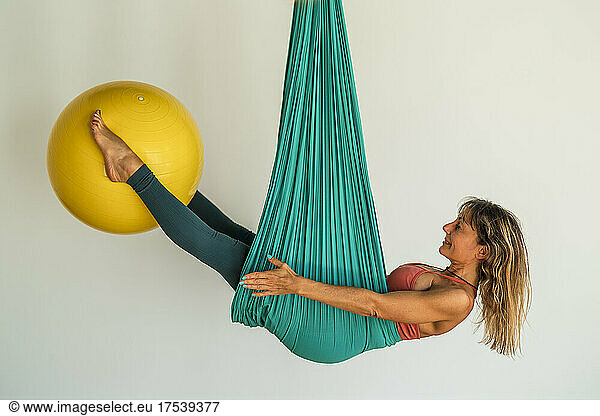Woman hanging on aerial silk holding fitness ball with legs in front of wall