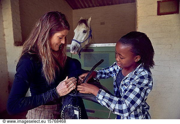 Woman helping girl prepare saddle in horse stables