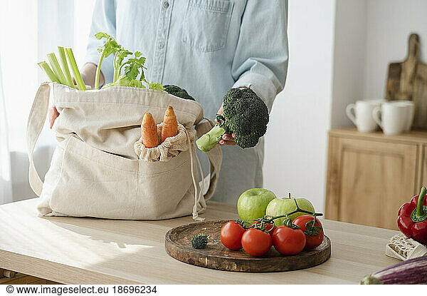 Woman holding broccoli by fresh fruits and vegetables on table at home