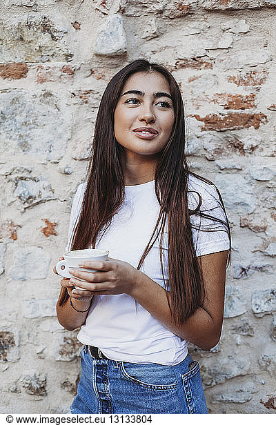 Woman holding cappuccino looking away while standing by old brick wall