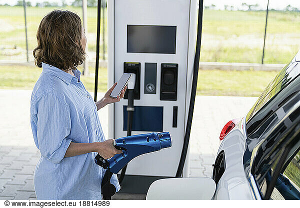 Woman holding car charger and smart phone next to electric car at station